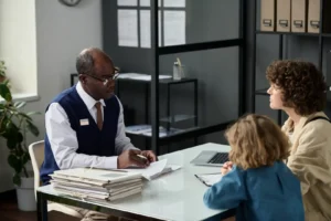 n older Black male advisor in glasses, sweater vest, and tie sitting at a white office desk reviewing paperwork across from a curly-haired mother and her young child.