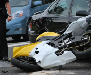 A damaged yellow and white sport motorcycle lying on its side on a city street following an accident with a black passenger car, with a law enforcement officer standing nearby to document the scene.