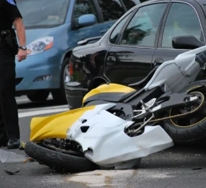 A damaged yellow and white sport motorcycle lying on its side on a city street following an accident with a black passenger car, with a law enforcement officer standing nearby to document the scene.