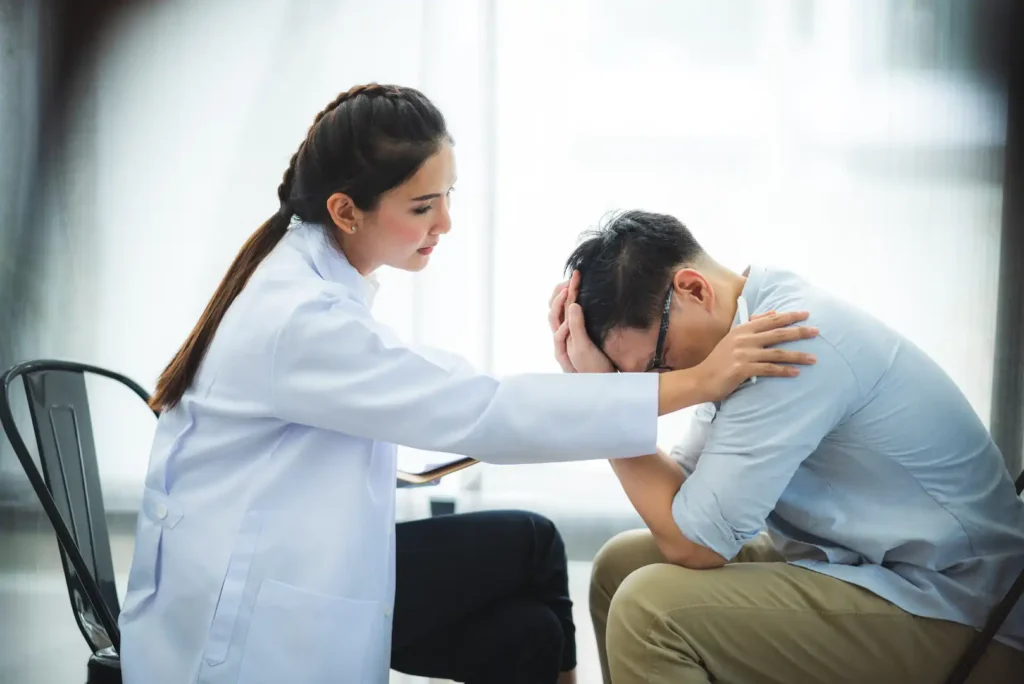 Side view of a female therapist or doctor in a white coat placing a supportive hand on the shoulder of a man who is leaning forward with his head in his hands during a counseling session.